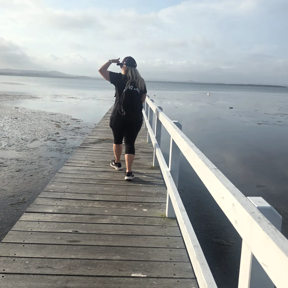 A woman walking out along a timber jetty over still water, shading her eyes as she looks to the horizon