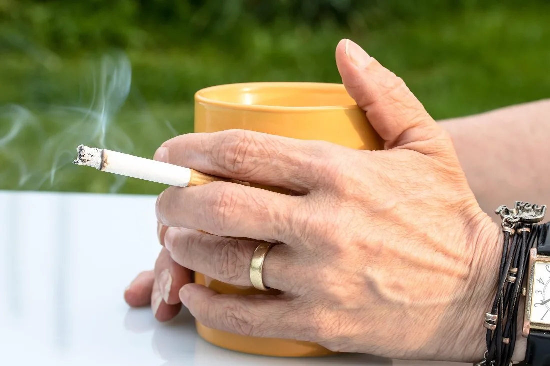 A hand holding a lit cigarette and a coffee mug